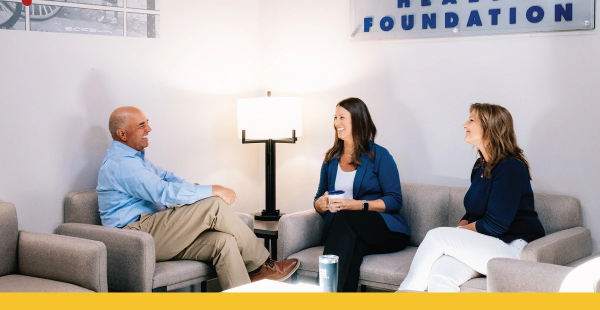 K21 staff seated and laughing together within their office. White walls, tan couches, and a yellow graphic bar at the bottom