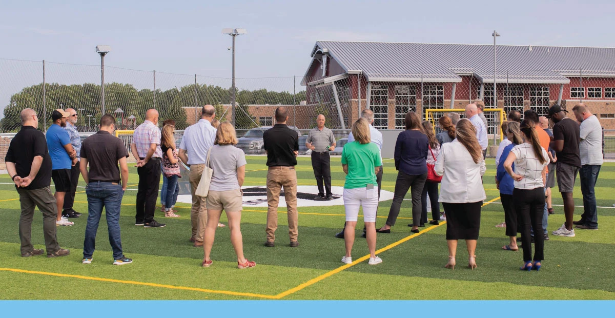Grand opening of the Kosciusko County YMCA soccer fields. Rich Haddad speaking to a large group surrounding him. Light blue graphic bar at the bottom.